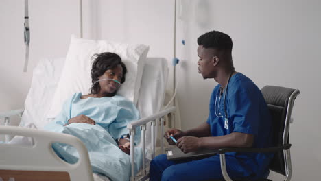 An-African-male-doctor-interviews-a-patient-lying-in-a-hospital-bed-with-an-oxygen-mask.-A-black-woman-lying-in-a-hospital-bed-describes-the-symptoms-to-the-doctor